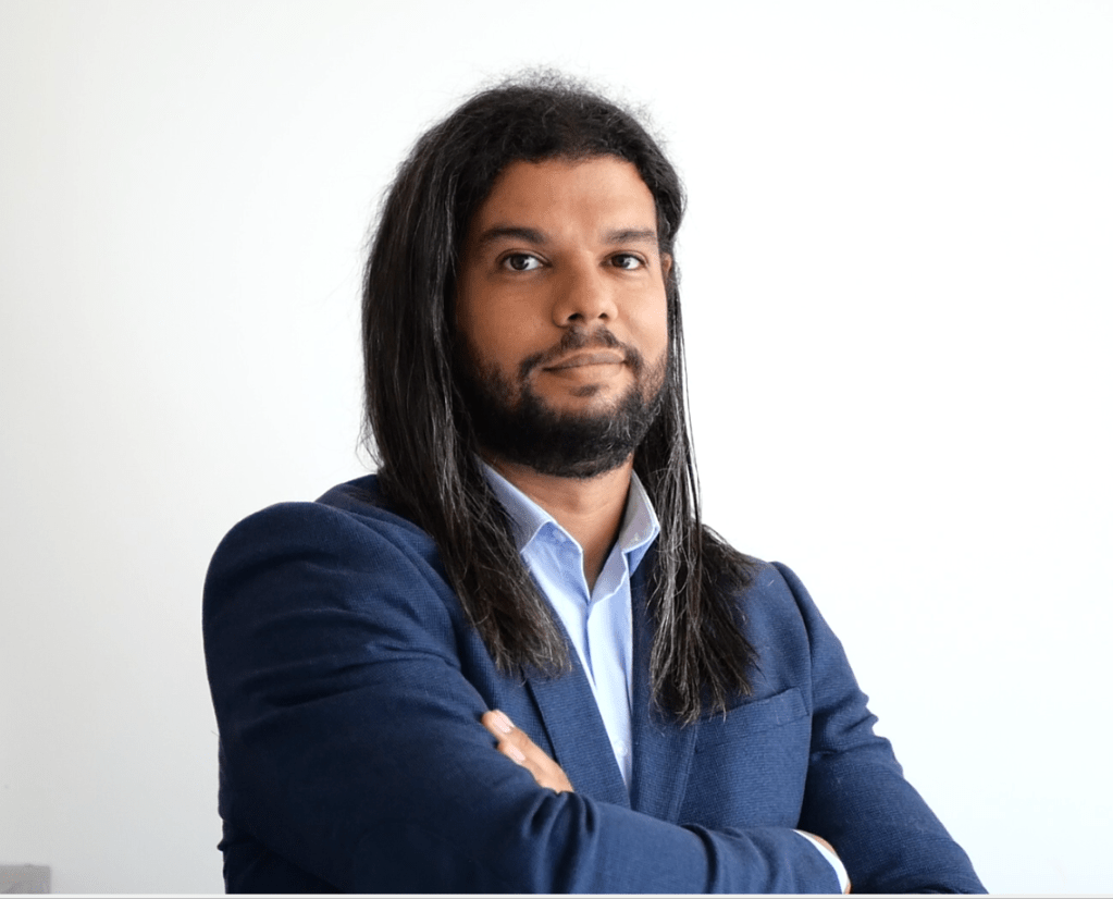 A man with long, dark hair and a dark beard, wearing a blue blazer and a light blue shirt. He has his hand under his chin ...